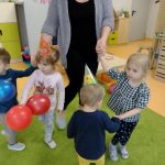 Dzieci razem z opiekunką trzymają się za ręce i tańczą w kółeczku na zielonym dywanie. Solenizantka stoi w środku i trzyma kolorowe baloniki.