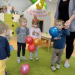 Dzieci stoją na zielonym dywanie i trzymają w rączkach kolorowe baloniki.