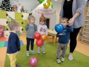 Dzieci stoją na zielonym dywanie i trzymają w rączkach kolorowe baloniki.