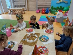Dzieci w kolorowych czapeczkach urodzinowych siedzi przy stoliku. Na stoliku kolorowe talerzyki i babeczki