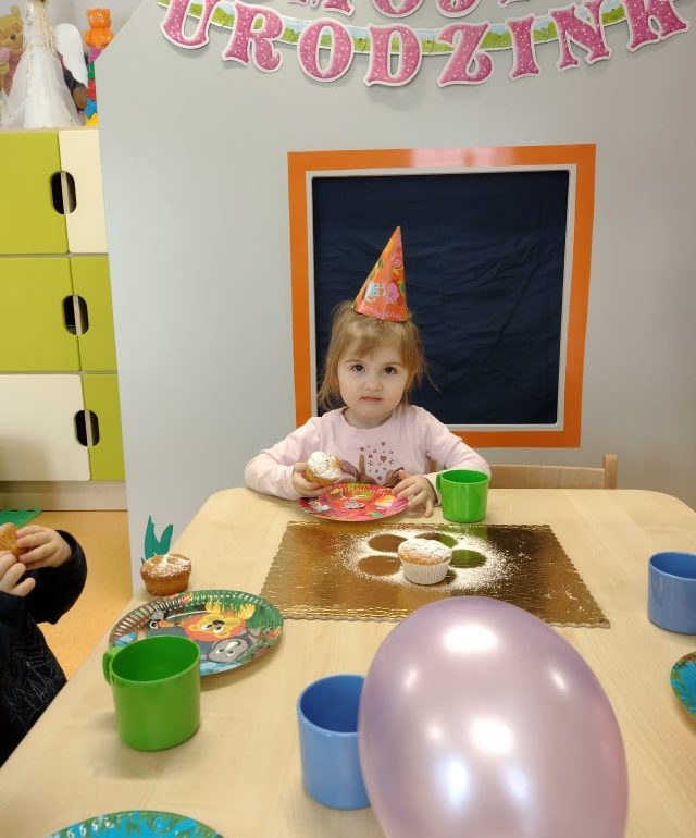 Dziewczynka siedzi przy stoliku, na głowie ma ubraną kolorową urodzinową czapeczkę. W rączkach trzyma babeczkę. W tle urodzinowy napis.