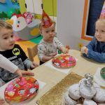 Dzieci siedzą przy stoliku w kolorowych czapeczkach urodzinowych. Na stoliku kolorowe papierowe talerzyki i babeczki posypane cukrem pudrem