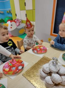 Dzieci siedzą przy stoliku w kolorowych czapeczkach urodzinowych. Na stoliku kolorowe papierowe talerzyki i babeczki posypane cukrem pudrem