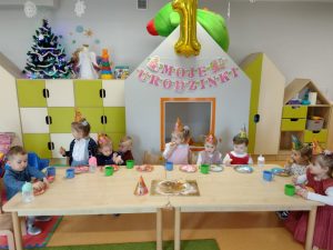 Dzieci w grupie siedzą przy stoliku. Na stoliku kolorowe talerzyki i kubeczki oraz tort z babeczek