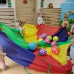 Dzieci w kole trzymają kolorową chustę, na której znajdują się kolorowe baloniki.