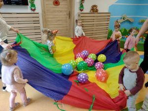 Dzieci w kole trzymają kolorową chustę, na której znajdują się kolorowe baloniki.