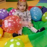 Dziewczynka z urodzinową czapeczką na głowie siedzi wśród balonów na kolorowej chuście.