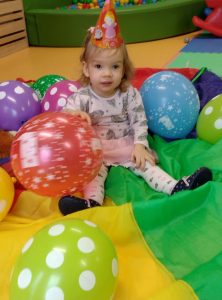 Dziewczynka z urodzinową czapeczką na głowie siedzi wśród balonów na kolorowej chuście.