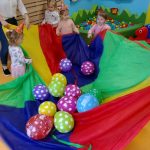 Dzieci trzymają w kole kolorową chustę z powrzucanymi na nią kolorowymi balonikami