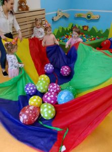 Dzieci trzymają w kole kolorową chustę z powrzucanymi na nią kolorowymi balonikami