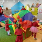 Dzieci ustawione w kole trzymają kolorową chustę z umieszczonymi na niej czerwonymi balonikami