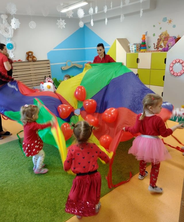 Dzieci ustawione w kole trzymają kolorową chustę z umieszczonymi na niej czerwonymi balonikami