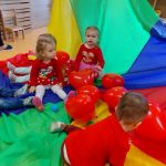 Grupa dzieci siedzi na kolorowej chuście wśród czerwonych baloników