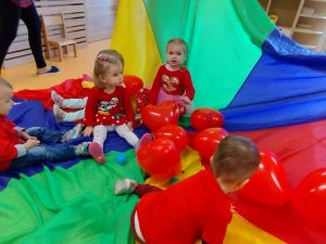 Grupa dzieci siedzi na kolorowej chuście wśród czerwonych baloników