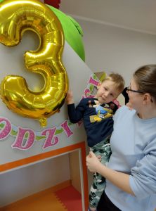Opiekunka trzyma na rękach solenizanta przy urodzinowym napisie i baloniku z cyfrą 3.