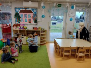 Dzieci z opiekunami obserwują postać ubraną w długi czarny płaszcz, czapkę z jasnego futra trzymającą sopel lodu