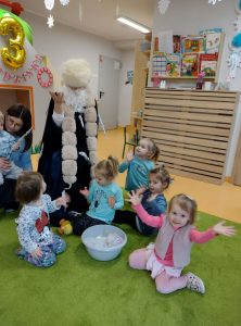 Wspólne zdjęcie z postacią w długim czarnym płaszczu. Dzieci siedzą na zielonym dywanie.. Postać stoi za nimi