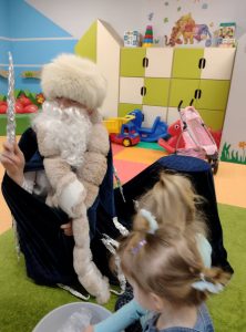 Postać ubrana w długi czarny płaszcz oraz futrzaną czapkę, w ręku trzyma sopel lodu. Postać ma długą białą brodę.