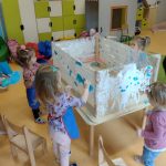Dzieci malują białą farbą abstrakcyjne plamy na rozwieszonej pomiędzy nogami stolika przezroczystej folii. Stolik jest położony do góry nogami na innym stoliku. Dzieci stoją wokół tego stolika.