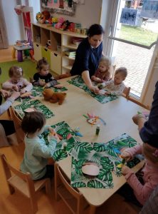 Dzieci siedzą przy stoliku. Na zielonej podkładce przyklejają kolorowe piórka, tworząc z nich pióropusz dla Indianinaa.