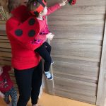Opiekunka ubrana w czerwoną bluzkę i czarne spodnie podnosi dziecko ubrane w strój biedronki. Dziecko pokazuje rączką przyklejoną z papieru biedronkę na drzwiach.