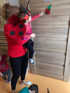Opiekunka ubrana w czerwoną bluzkę i czarne spodnie podnosi dziecko ubrane w strój biedronki. Dziecko pokazuje rączką przyklejoną z papieru biedronkę na drzwiach.