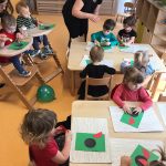 Dzieci siedzą przy stoliku i przyklejają na zielonej kartce papieru czerwoną biedronkę z kółeczek.