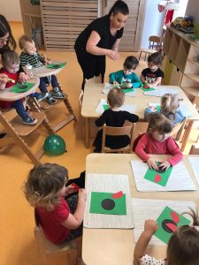 Dzieci siedzą przy stoliku i przyklejają na zielonej kartce papieru czerwoną biedronkę z kółeczek.