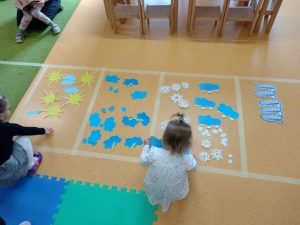 Dzieci układają symbole zjawisk atmosferycznych na wyznaczonych kwadratach na podłodze.
