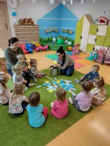 Dzieci siedzą w okręgu na zielonym dywanie. Po środku znajdują się różne symbole zjawisk atmosferycznych. Opiekunka pokazuje dzieciom jak wygląda marcowa zupa, trzyma w ręce papierowe słoneczko i wkłada je do garnka.