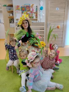 Pani Wiosna siedzi w śród dzieci ubrana w piękną sukienkę w kwiaty, długi zielony płaszcz oraz piękny słomkowy kapelusz udekorowany kwiatami, w ręce trzyma wiklinowy koszyk wypełniony kwiatami.