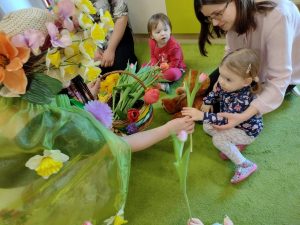 Dzieci siedzą po turecku na zielonym dywanie. Pani Wiosna rozdaje dzieciom czerwone tulipany.