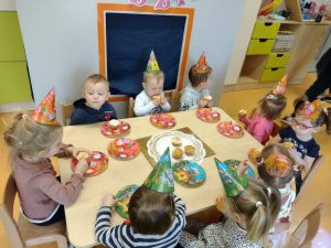 Dzieci siedzą przy stoliku i częstują się mufinkami. Na główkach mają ubrane kolorowe czapeczki. W tle witać napis urodzinki i dużą złotą cyfrę 2.