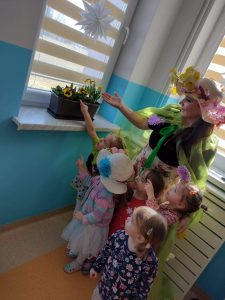 Dzieci wraz z Panią Wiosną podziwiają na oknie w szatni kolorowe kwiatuszki posadzone w doniczce przez dzieci na zajęciach.