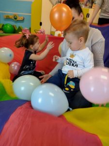Opiekunka siedzi na kolorowej chuście i trzyma chłopca na kolanach . Dziewczynka z tylu podrzuca kolorowe baloniki do góry.