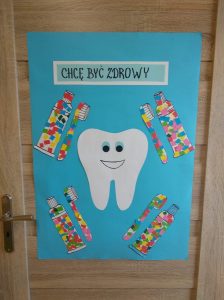 Na drzwiach wiski duży niebieski karton z napisem "Chcę być zdrowy" na środku plakatu znajduje się biały duży ząb z uśmiechniętą miną, dookoła zęba znajdują się kolorowe pasty i szczoteczki.