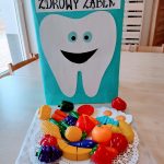 Na stoliku stoi niebieskie pudełko z naklejonym dużym białym ząbkiem z uśmiechniętą buzią i napisem "Zdrowy ząbek". Przed pudełkiem na białej serwetce leżą zabawkowe owoce i warzywa.