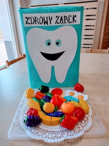 Na stoliku stoi niebieskie pudełko z naklejonym dużym białym ząbkiem z uśmiechniętą buzią i napisem "Zdrowy ząbek". Przed pudełkiem na białej serwetce leżą zabawkowe owoce i warzywa.