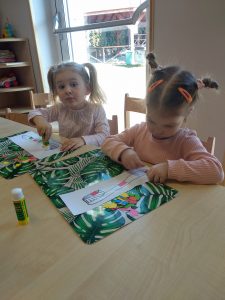 Dzieci siedzą przy stoliku. Na zielonej podkładce wyklejają kolorowymi kwadratami narysowaną szczoteczkę i pastę.