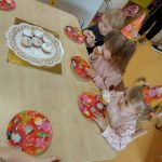 Dzieci siedzą w urodzinowych czapeczkach przy stoliku i częstują się urodzinowymi babeczkami.