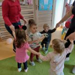 Dzieci trzymają się za rączki i tańczą w kółeczku. W środku stoi chłopczyk z balonikiem w rączkach i tańczy razem z dziećmi.