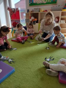 Dzieci siedzą po turecku na zielonym dywanie. Opiekunka pokazuje dzieciom gniazdo ptaka. Przed każdym dzieckiem leży papierowa jaskółka.