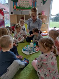 Dzieci siedzą po turecku na dywanie w dużym kole, Na środku stoi taca z mlecznymi produktami. Opiekunka pokazuje dzieciom mleczne produkty. Dzieci z wielkim zainteresowaniem słuchają i oglądają produkty.
