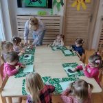 Dzieci siedzą przy stoliku na zielonej podkładce przyklejają z gotowych elementów czarną krówkę. W ile widać telewizor z wyświetloną pracą plastyczną pt. "krówka".