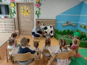 Dzieci siedzą na krzesełkach i patrzą jak opiekunka pokazuje makietę krowy.