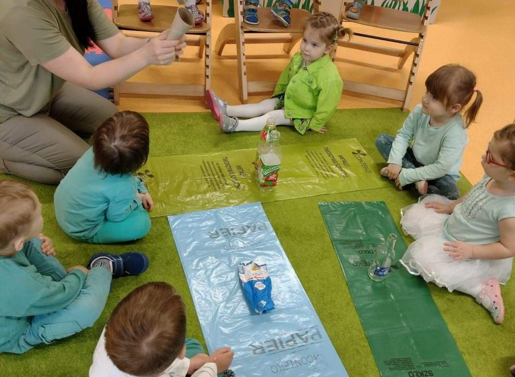 Dzieci siedzą na zielonym dywanie i w drewnianych krzesełkach. Na środku opiekunka pokazuje jak należy segregować śmieci. W dłoniach trzyma przykładowe śmieci, które układa na rozłożonych workach do segregacji. Dzieci patrzą z wielkim zainteresowaniem.