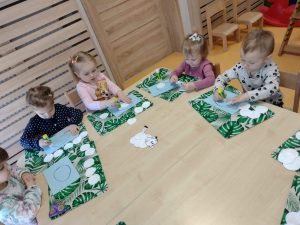 Dzieci siedzą przy stoliku , na zielonej podkładce wyklejają baranka z białych wacików kosmetycznych.