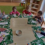 Dzieci siedzą przy stoliku, na zielonej podkładce wyklejają kolorowymi kółeczkami wielkanocne jajko.