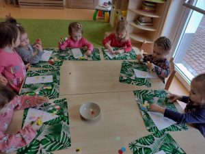Dzieci siedzą przy stoliku, na zielonej podkładce wyklejają kolorowymi kółeczkami wielkanocne jajko.