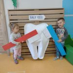 Dzieci stoją koło dużego białego zęba w rączkach trzymają duże papierowe szczoteczki w kolorze niebieskim i czerwonym.